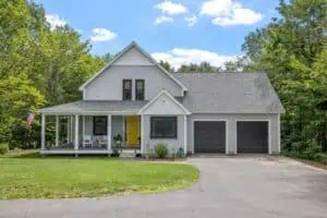 Gray house with pitched roof, porch with yellow door, two-car garage. Consider professional roofing maintenance.