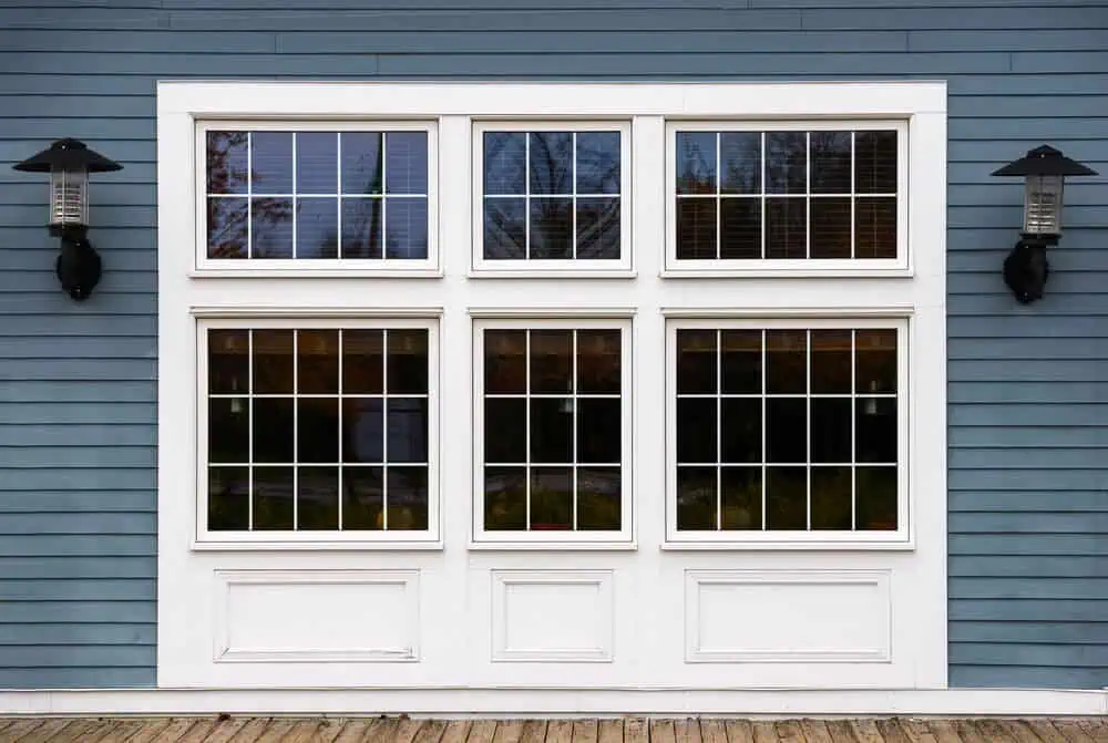 A large white-framed window with six panes and two black wall-mounted lanterns adorn the blue wooden exterior, showcasing the classic elegance while highlighting the impressive lifespan of replacement windows in Michigan.