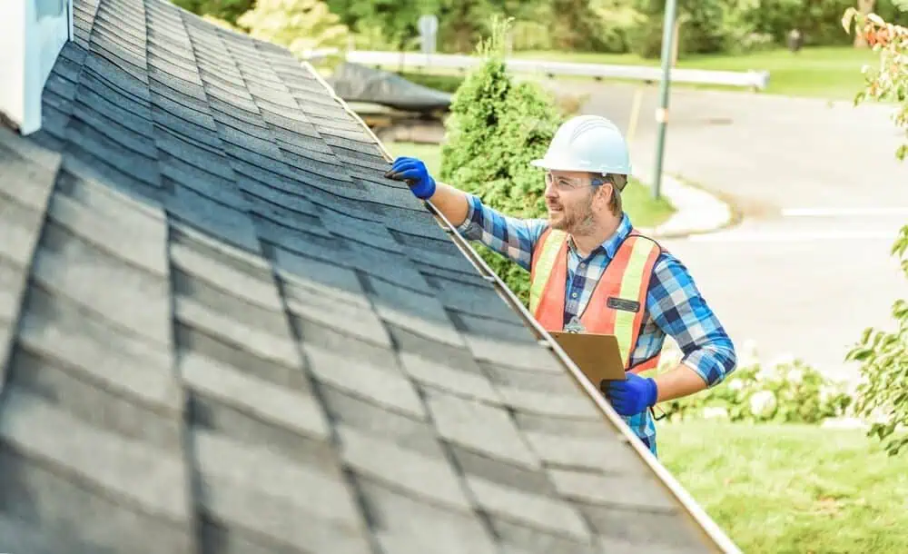 Roof inspector in a hard hat and safety vest checking shingles on the eave of a roof.