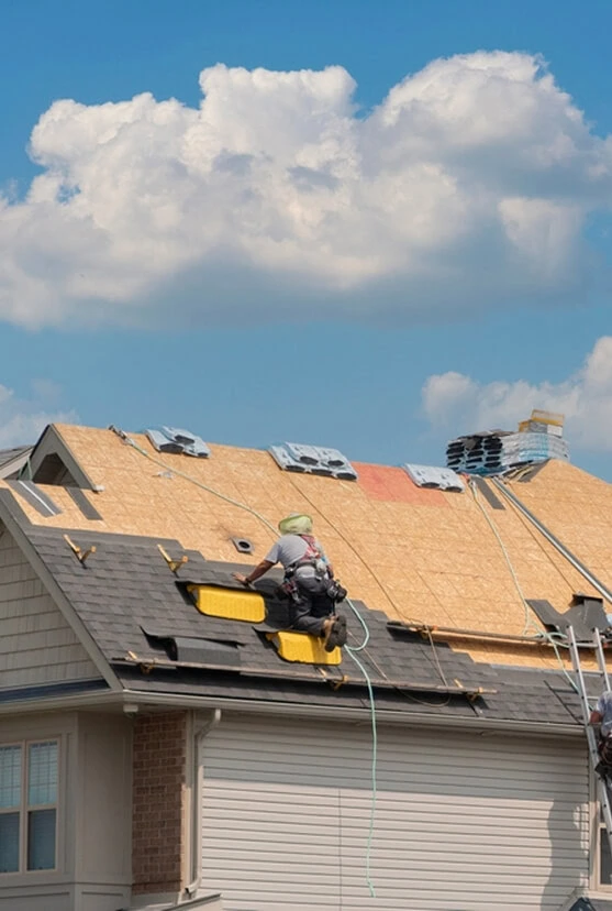 Michigan roofing services contractor repairing shingles with tools, gutters, and safety gear visible under blue sky.